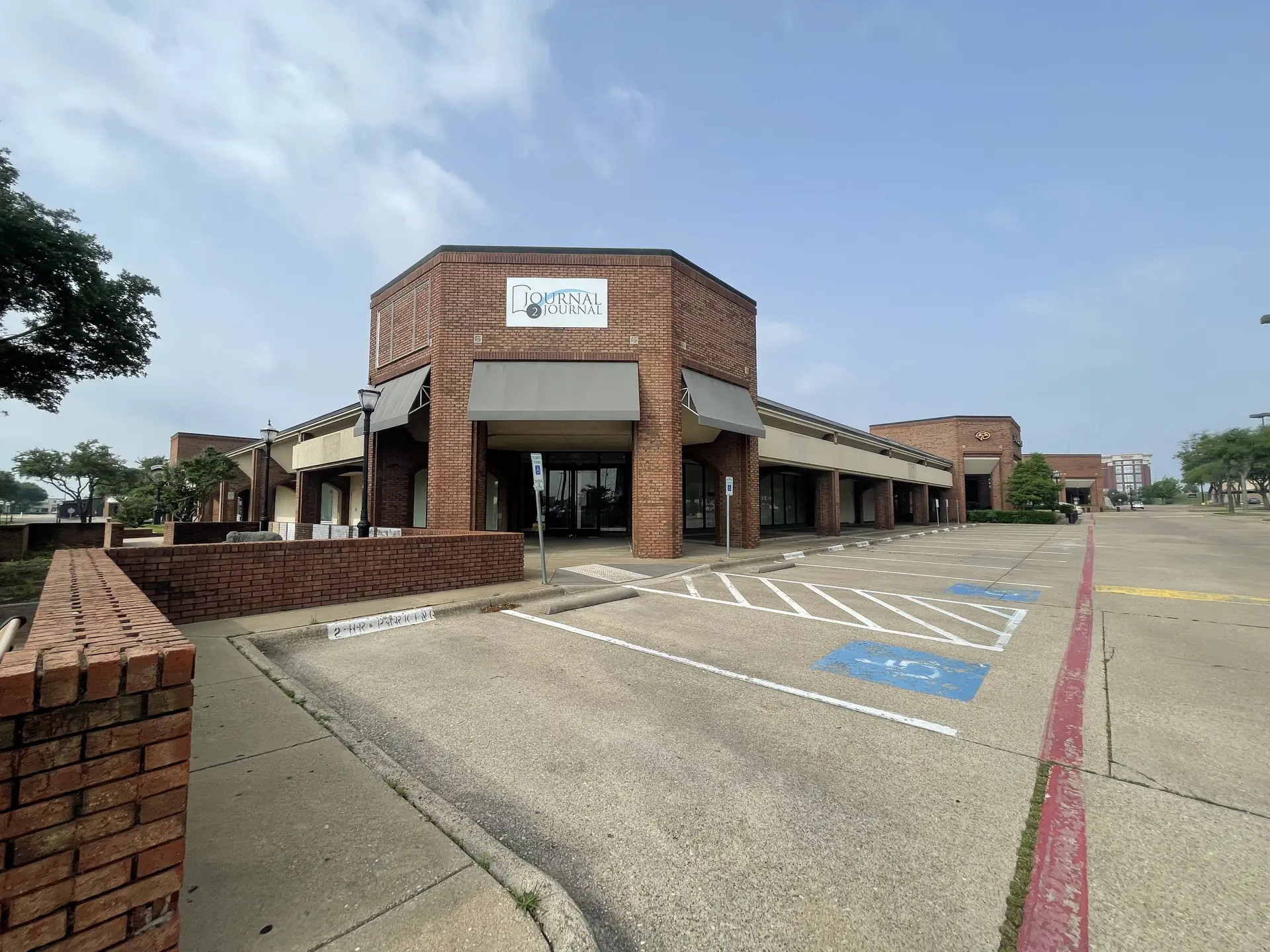 Exterior view of a Journal 2 Journal brick building in a shopping center with an empty parking lot, grey awnings, and a blue sky with light clouds.