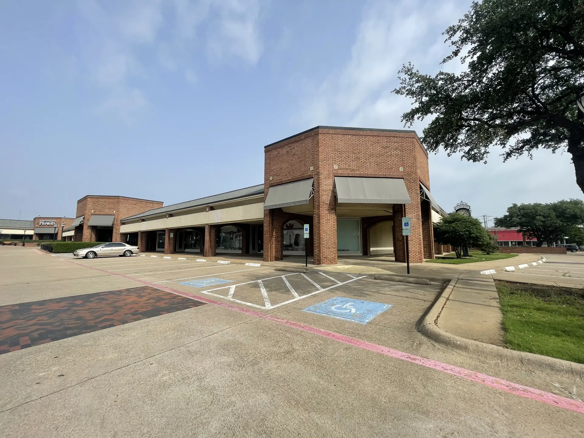 Exterior view of an older red brick retail storefront with grey awnings, an empty parking lot with blue handicap stalls, and a large tree under a hazy sky.