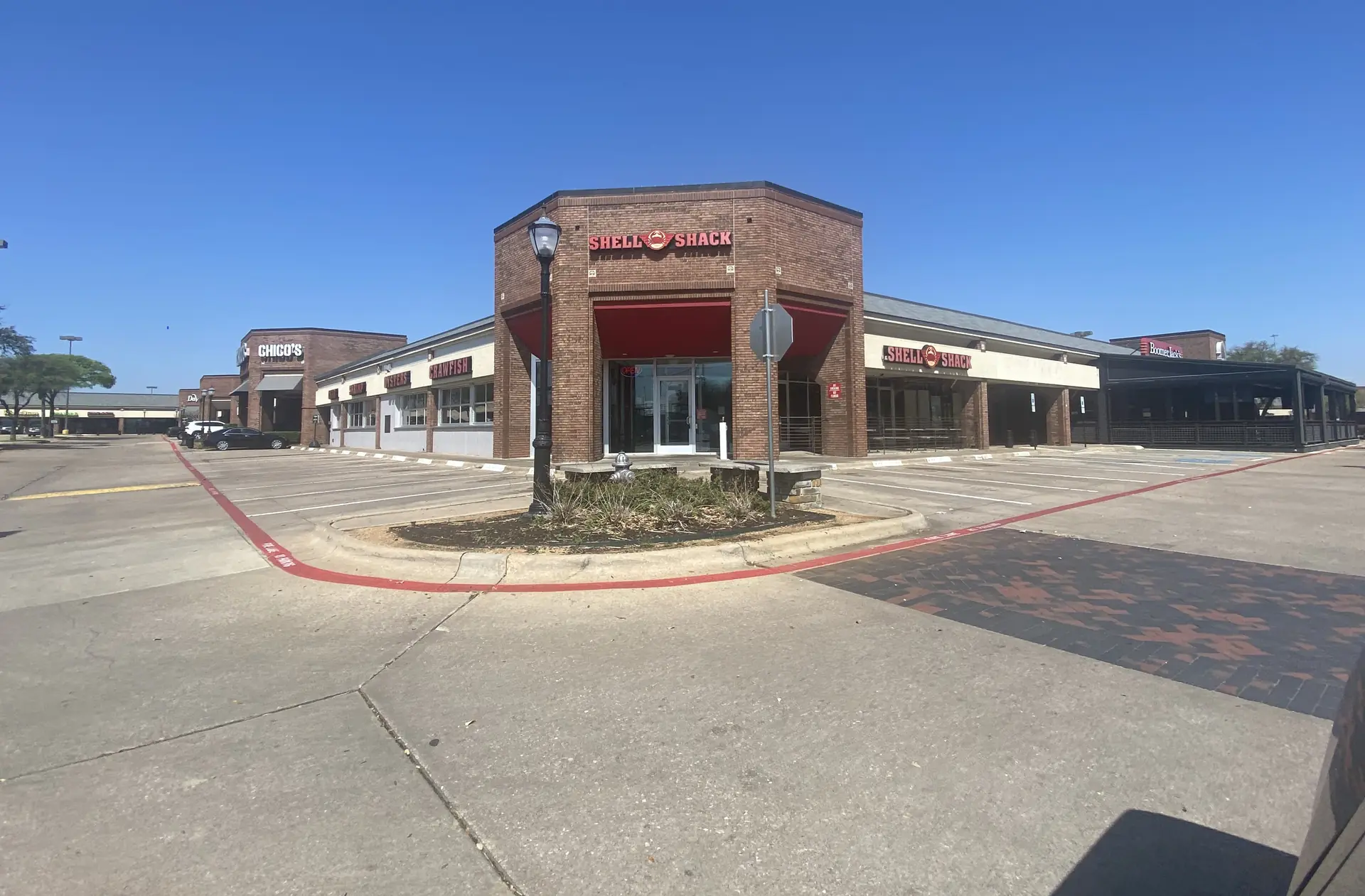 Exterior view of "Shell Shack" in a traditional red brick retail building with an empty parking lot, a black lamppost, and a clear, bright blue sky.