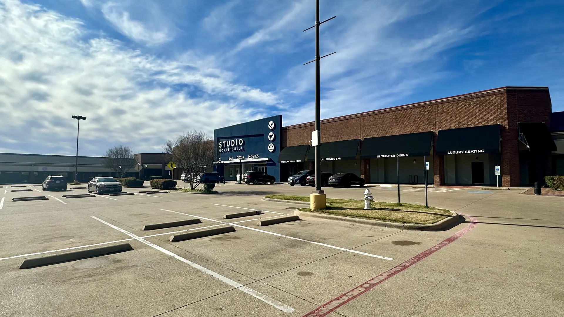 Exterior of a brick building featuring a blue "Studio Movie Grill" entrance with black awnings and "Luxury Seating" signs, facing a large, mostly empty parking lot under a bright, cloudy sky.